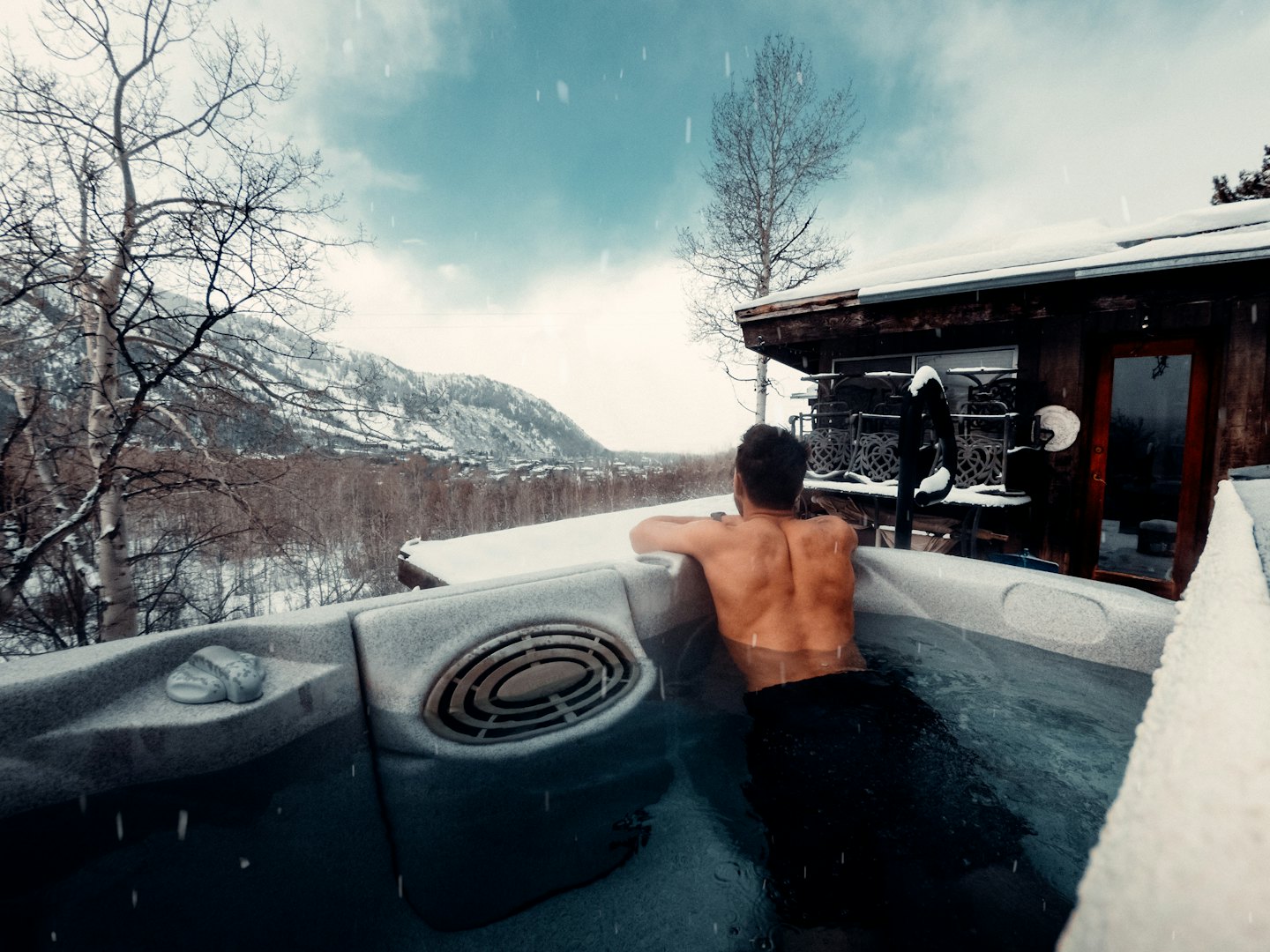 Man soaks in a hot tub overlooking snowy mountains.